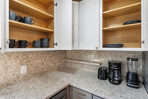 A kitchen with granite countertops, a coffee maker, a blender, and open cabinets with mugs and plates.
