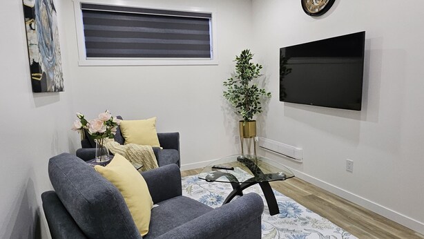 A living room with a grey sofa, a flat-screen TV, a window with blinds, a potted plant, and a floral rug. - Xquisithomes: More than a stay, it's an experience (Saskatoon)