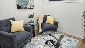 A modern living room with two grey sofas, a glass coffee table, a floral rug, and a large abstract painting on the wall. - Xquisithomes: More than a stay, it's an experience (Saskatoon)