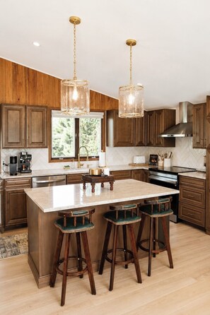 A kitchen with wooden cabinets, a large island with stools, pendant lights, and a window with a view. - Luxury Big Sky Golf Course Retreat with Hot Tub (Big Sky)