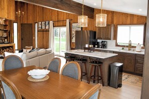 A modern kitchen with wooden cabinetry, a central island, bar stools, and a dining table with chairs. - Luxury Big Sky Golf Course Retreat with Hot Tub (Big Sky)