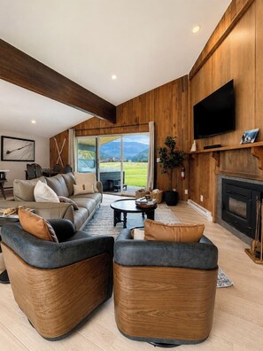 A modern living room with a fireplace, a large flat-screen TV, and a view of the outdoors. - Luxury Big Sky Golf Course Retreat with Hot Tub (Big Sky)
