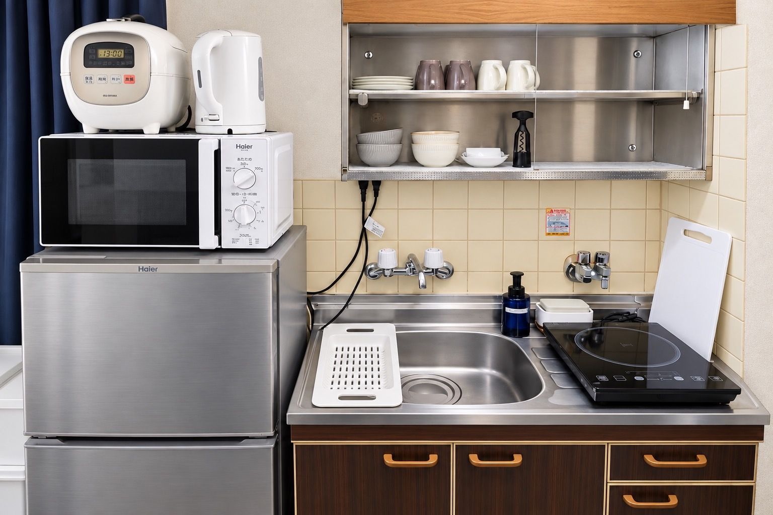 A compact kitchen with a microwave, toaster, kettle, and a sink with a cutting board.
