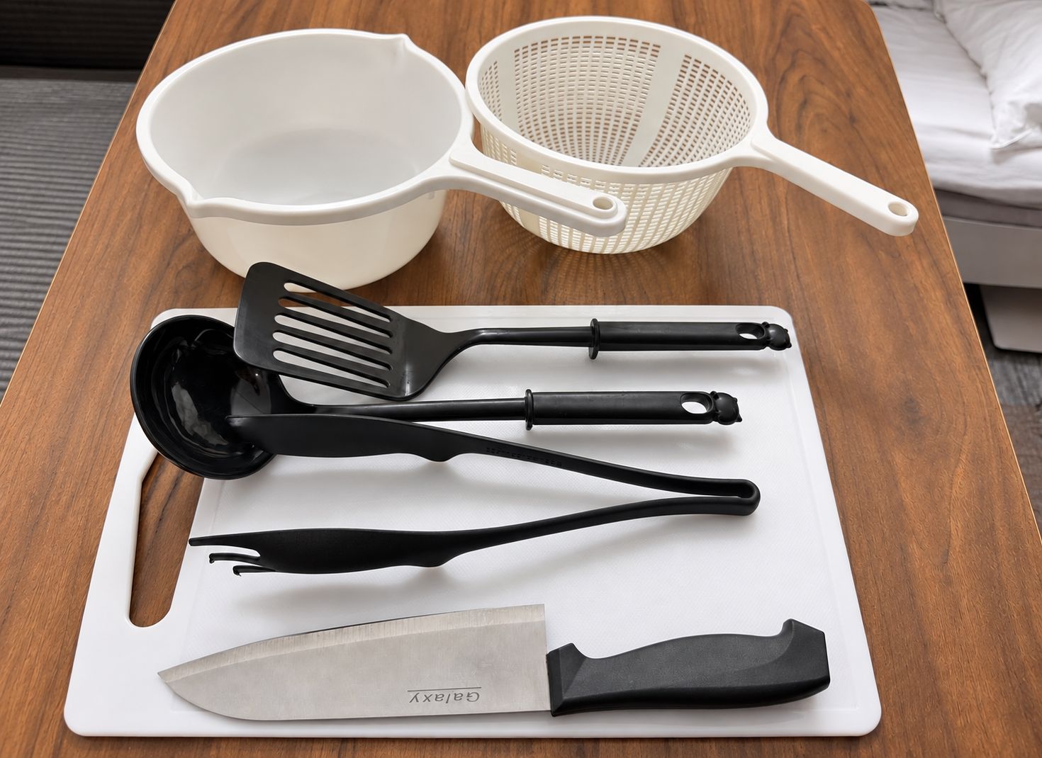 A kitchen counter with a white cutting board, a black spatula, a black ladle, a black strainer, and a white bowl.