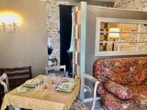 A dining area with a table set for two, a floral sofa, and a stone wall. - Les Fleurs De Lys, Suites&Appart , Entrée et Terrasse Privée Proche de la Mer (Saint-Georges-d'Oléron)