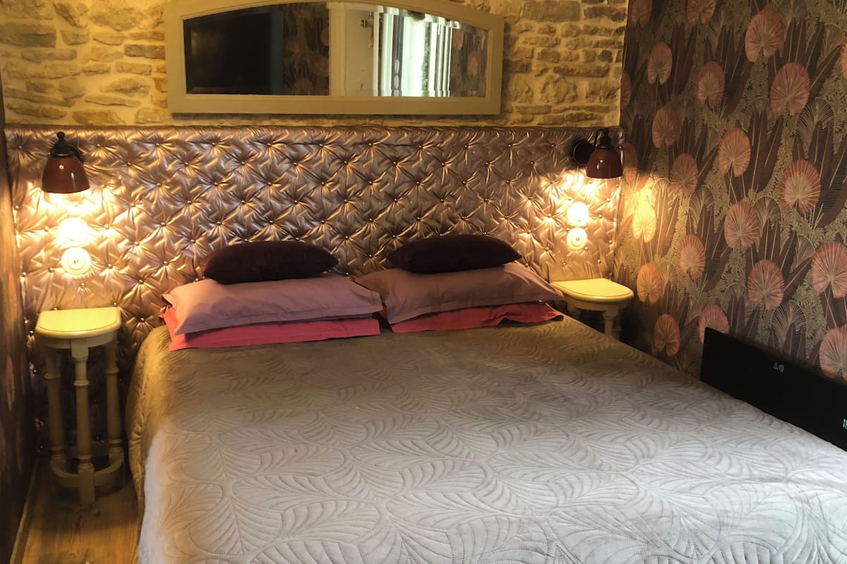 Un lit avec une tête de lit capitonnée, deux lampes de chevet, un miroir au-dessus de la tête de lit et un papier peint à motifs floraux sur le mur de droite.