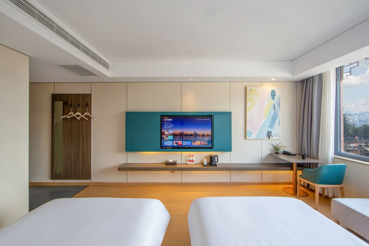 Habitación de hotel con dos camas, televisión, escritorio, silla, ventana con vista a la ciudad y perchero.