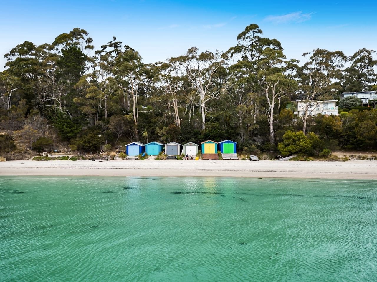 A beach with clear turquoise water, a row of colorful beach huts, and a forested area in the background.