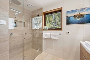 Un baño moderno con ducha envuelta en vidrio, una ventana con vista a árboles y un cuadro de un barco en un lago.