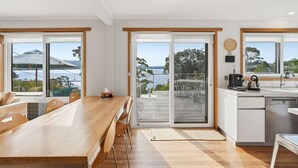 Ett modernt kök med utsikt över havet, en matplats med ett träbord och en skjutdörr i glas som leder ut till en uteplats.
