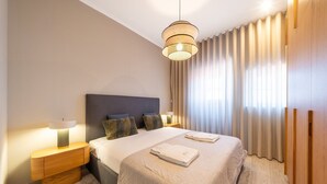 Et moderne soveværelse med en stor seng, to sengeborde, en hængende lampe og udsigt udad gennem lette gardiner.