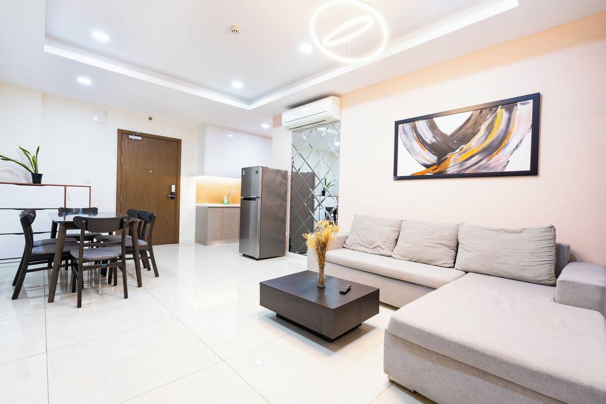A modern living room with a sofa, coffee table, dining table with chairs, and a kitchen area with a refrigerator.