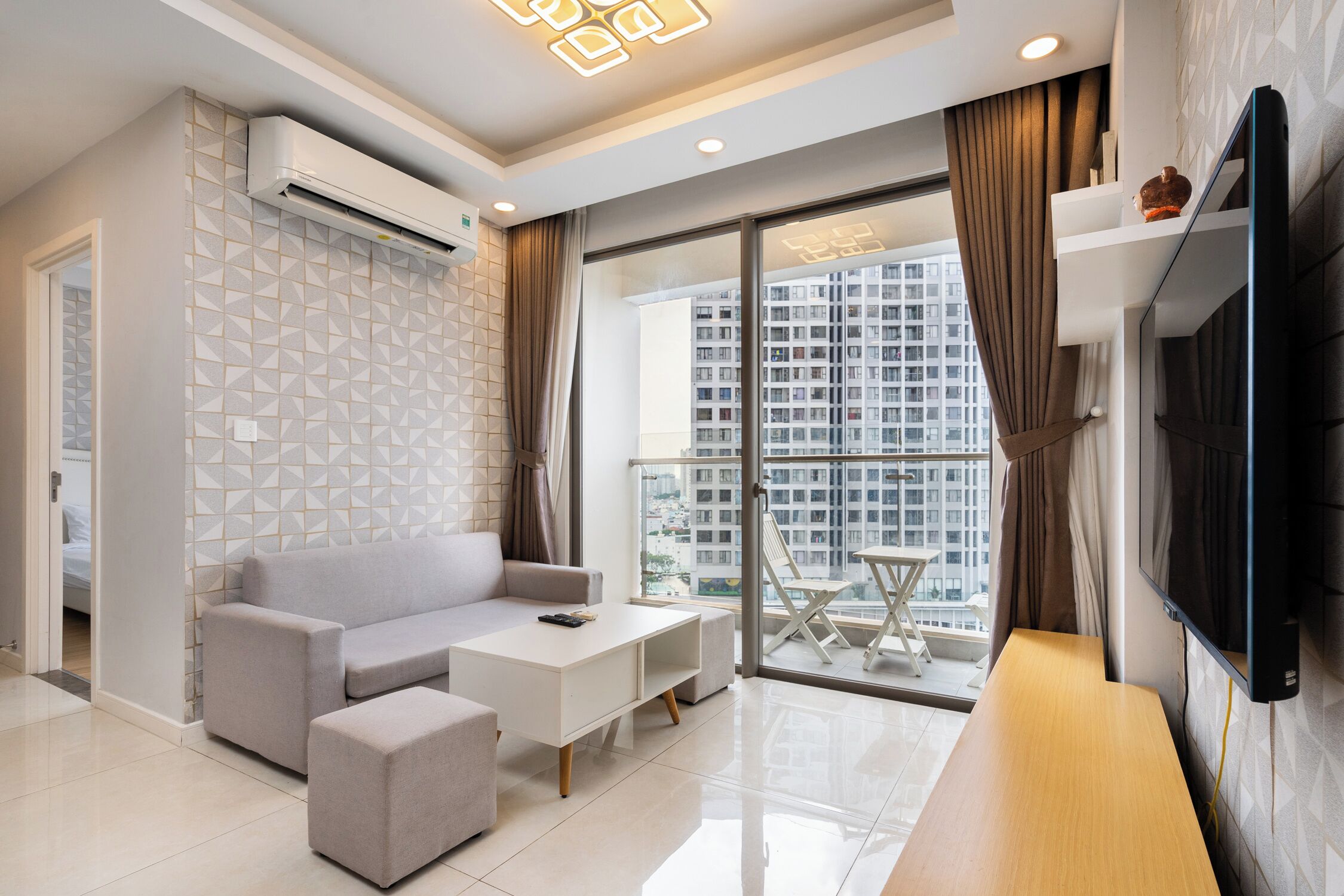 Una sala de estar moderna con sofá, mesa de centro y televisor, ventanas amplias con cortinas, y un balcón con mesa y sillas.