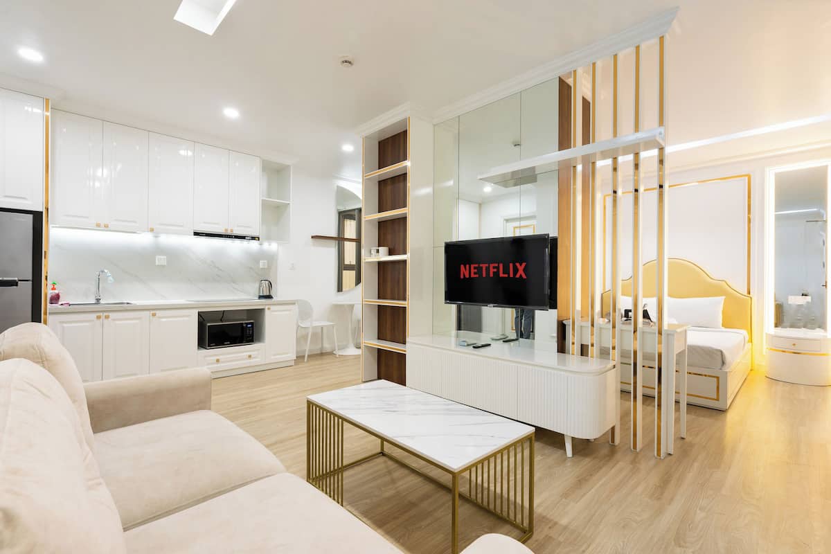 A modern living room with a sofa, a coffee table, a TV, a kitchen area, and a bedroom with a bed and nightstand.
