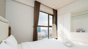 Habitación de hotel con cama, ventana con cortinas, vista a la ciudad, un espejo y repisas con objetos.