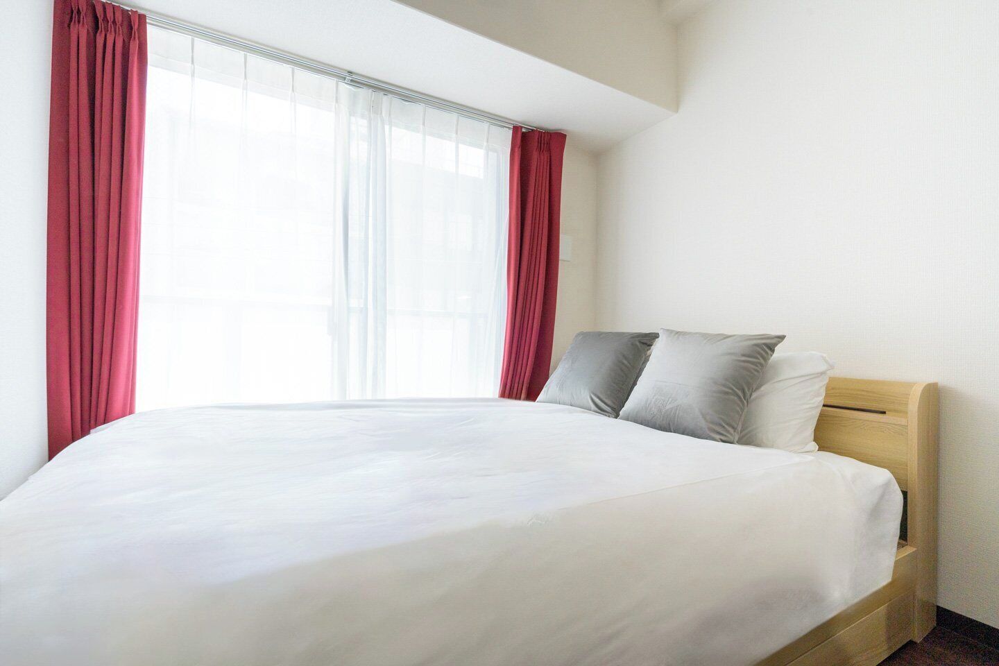Una cama individual con cabecera de madera, ropa de cama blanca y cortinas rojas sobre una pared blanca.