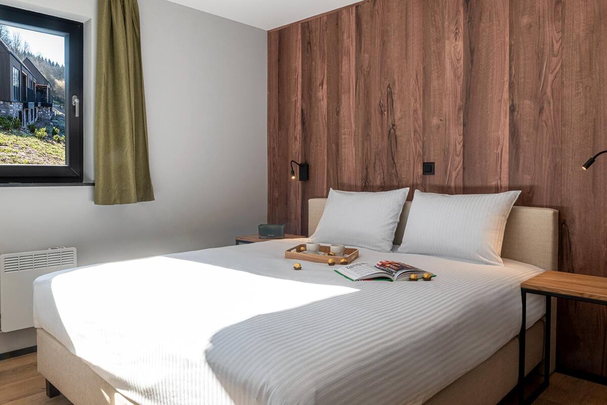 A bedroom with a wooden headboard, a window with a curtain, a bedside table with a lamp, and a bed with pillows and a tray of items.