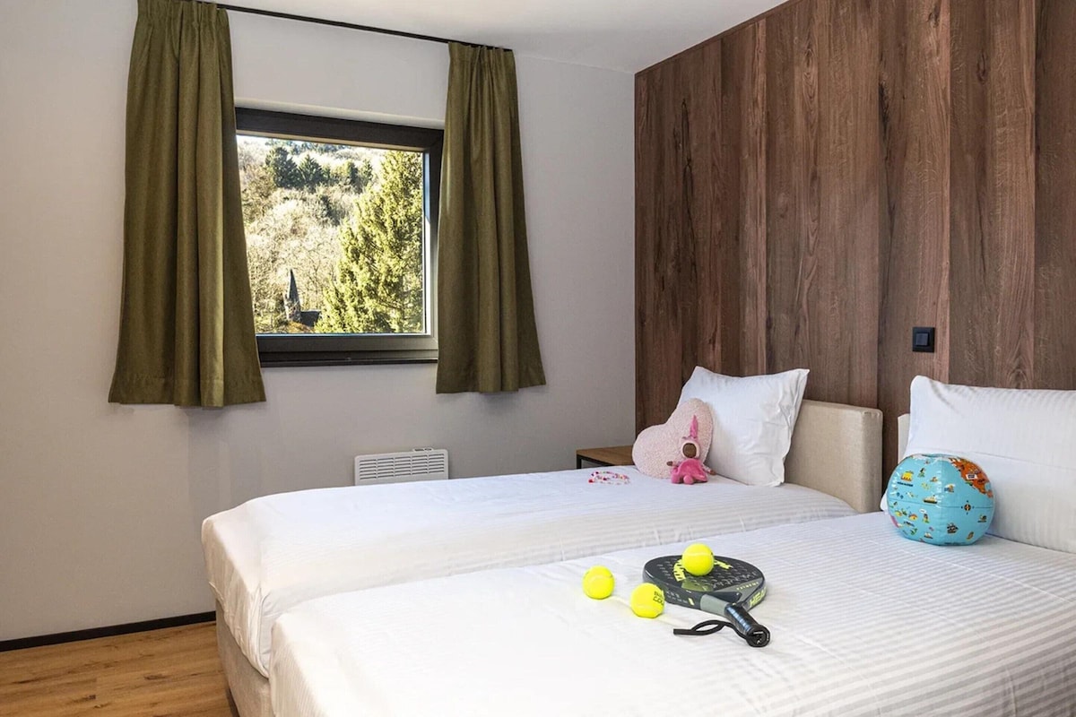 A bedroom with a large bed, a window with curtains, a wooden wall, and a view of trees outside.