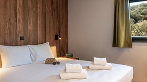 A hotel room with a wooden headboard, a bed with white linens, a bedside table with a lamp, and a window with a view of trees.