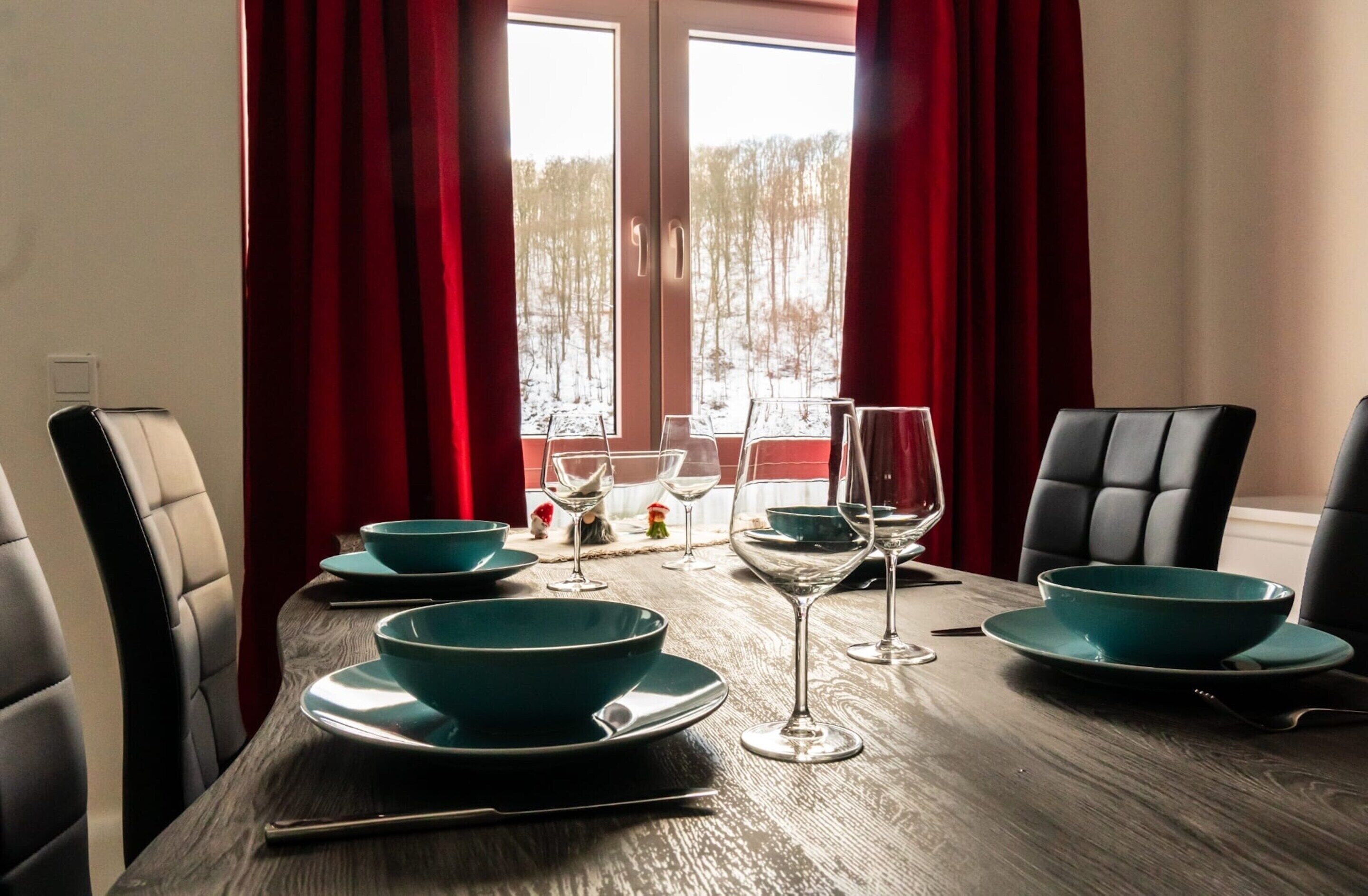 Une table à manger dressée avec des bols turquoise et des verres à vin, des rideaux rouges et une vue sur un paysage enneigé par la fenêtre.