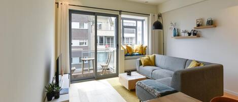 Un appartement moderne avec un canapé gris, une table à manger en bois et un balcon agrémenté de coussins jaunes.