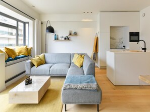 A modern living room with a grey sectional sofa, a wooden coffee table, and a kitchen area with a white countertop and black faucet. - Sunny Escape at Albert Beach (Knokke-Heist)