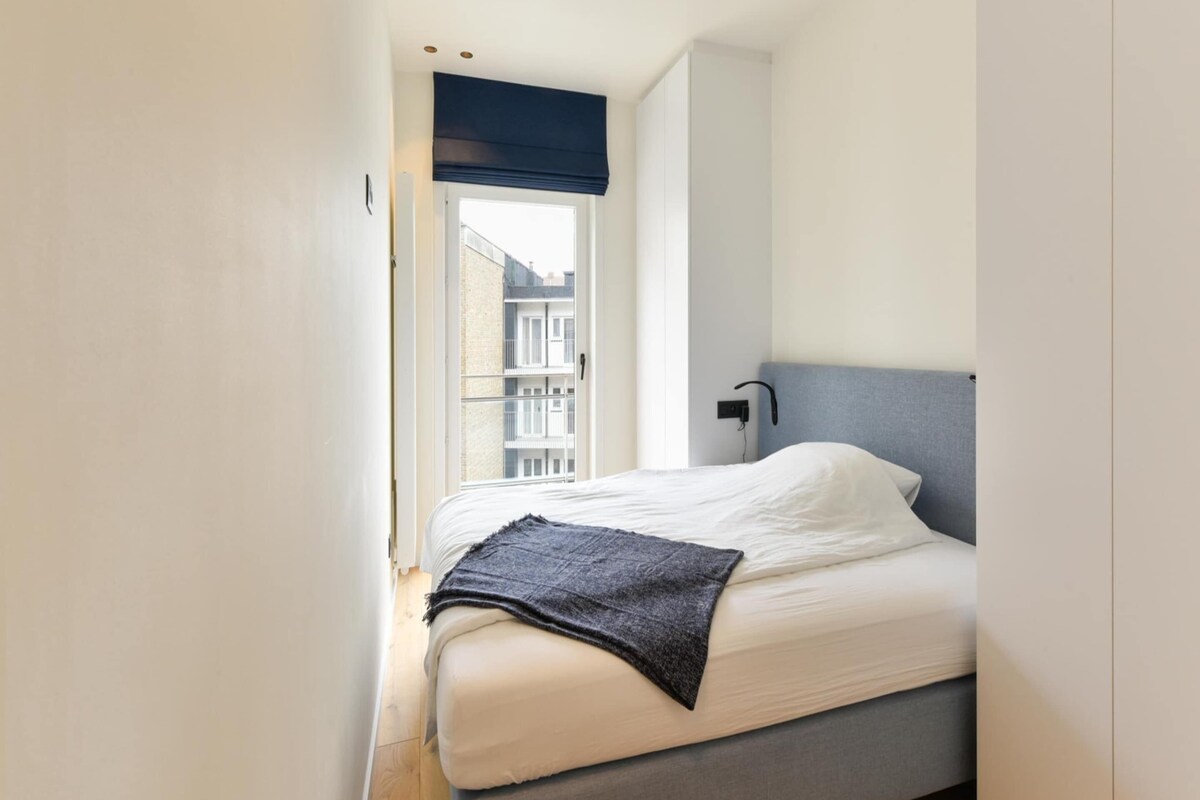 A bedroom with a bed, a window with a view of buildings, a bedside table, and a wardrobe.
