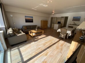 A spacious living area with a sofa, coffee table, and dining table. There is a kitchen area with a refrigerator and a microwave. The room has wooden flooring and large windows allowing natural light. - Sunny Escape at Albert Beach (Knokke-Heist)