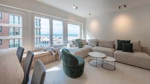 A modern living room with a sofa, two round coffee tables, and a green armchair. Large windows offer a view of the city and the sea. - Modern Seaside Escape (Knokke-Heist)