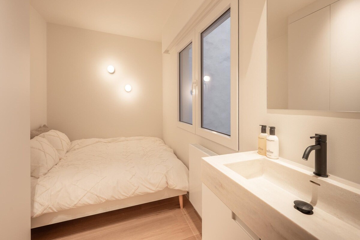 A bedroom with a bed, a window, a bathroom with a sink and mirror, and a nightstand with toiletries.