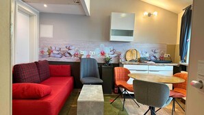 Un espacio habitable reducido con un sofá rojo, un sillón gris y sillas naranjas. Una pequeña zona de cocina con un lavabo y un muro decorativo.