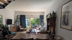 Un vaste espace de vie comprenant une table à manger en bois, des chaises et un canapé. On trouve une télévision sur le côté gauche et une bibliothèque agrémentée d'objets décoratifs.