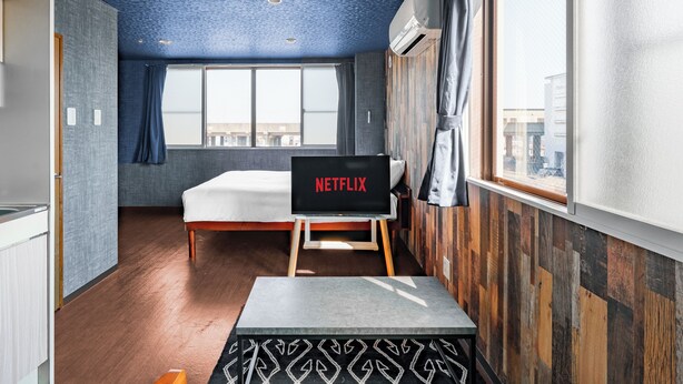 Una camera moderna con un letto, un tavolino, uno schermo TV che mostra 'NETFLIX' e una finestra con tende.