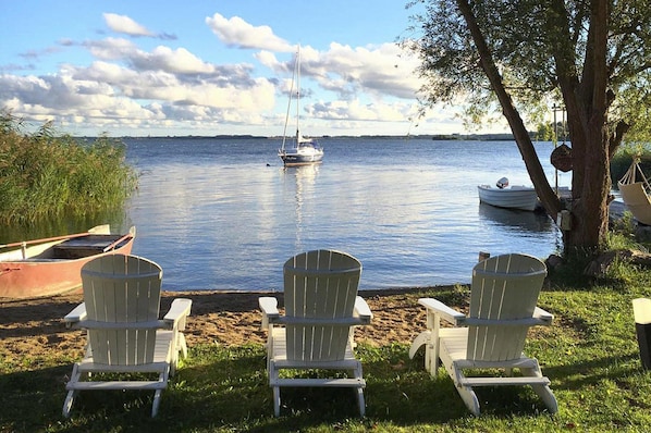 A lakeside view with three Adirondack chairs facing the water, a sailboat, and a rowboat docked near the shore. - Ferienhaus mit Kamin am Achterwasser in Lutow (Luetow)