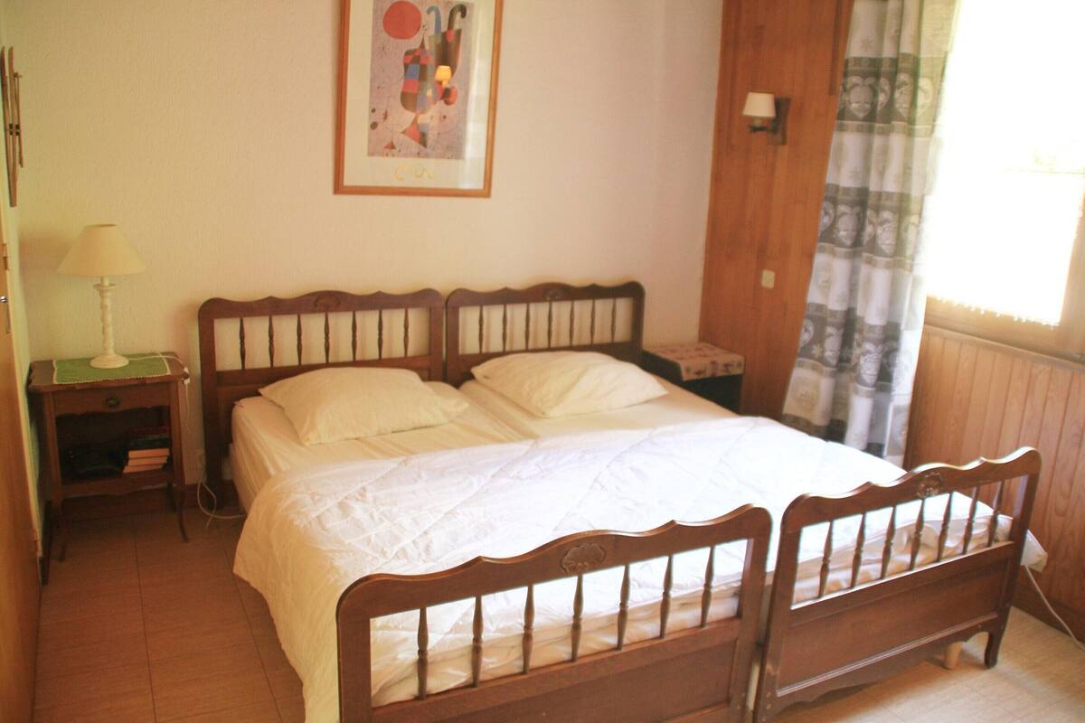 Une chambre à coucher avec un lit en bois, une table de chevet avec une lampe, un tableau accroché au mur et une fenêtre avec des rideaux.