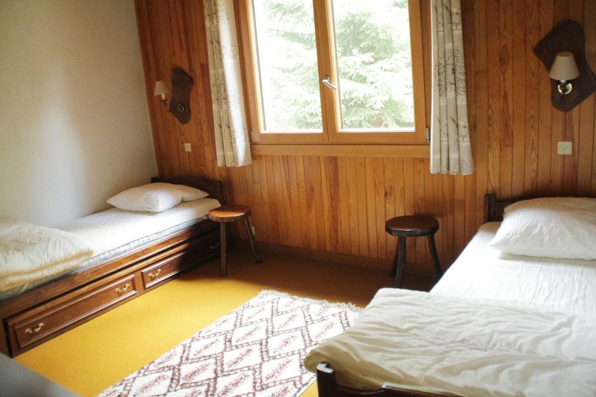 Une chambre avec deux lits, un plancher en bois, une fenêtre avec des rideaux et un petit tabouret.