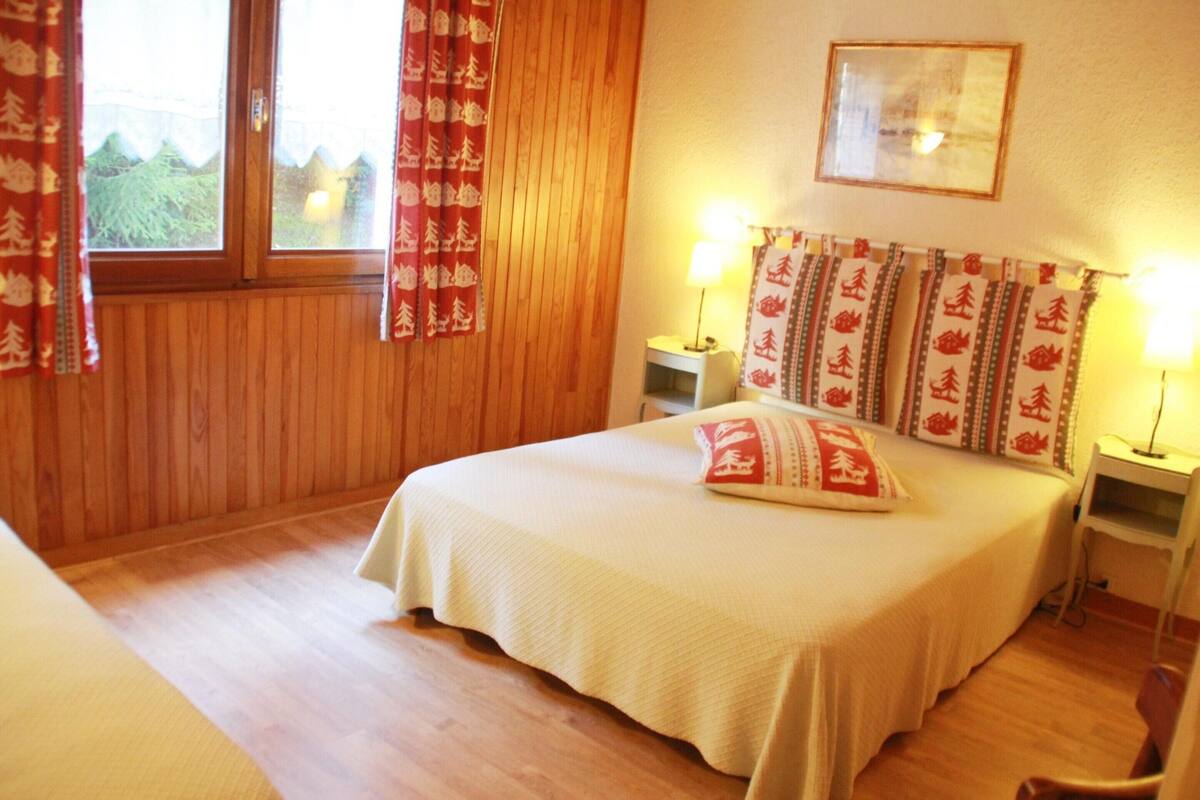 Une chambre avec un plancher en bois, un lit recouvert d’une courtepointe blanche, deux tables de chevet avec des lampes, et des rideaux à motifs rouges et blancs.