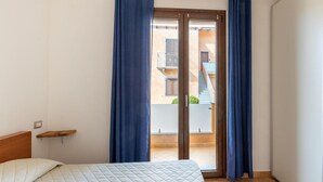 Quarto com cama, cabeceira de madeira, janela com cortinas azuis e vista para um edifício.
