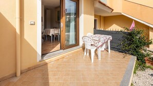 Um pátio com mesa e cadeiras, uma porta de vidro deslizante que dá para um espaço interno e uma pequena área de jardim com uma planta.