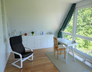 Une pièce avec un bureau blanc, une chaise noire, une chaise en bois, une armoire blanche et une fenêtre donnant sur des arbres.