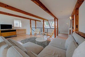 A spacious living room with a high ceiling, wooden beams, a large sofa, a TV, and a balcony with chairs.