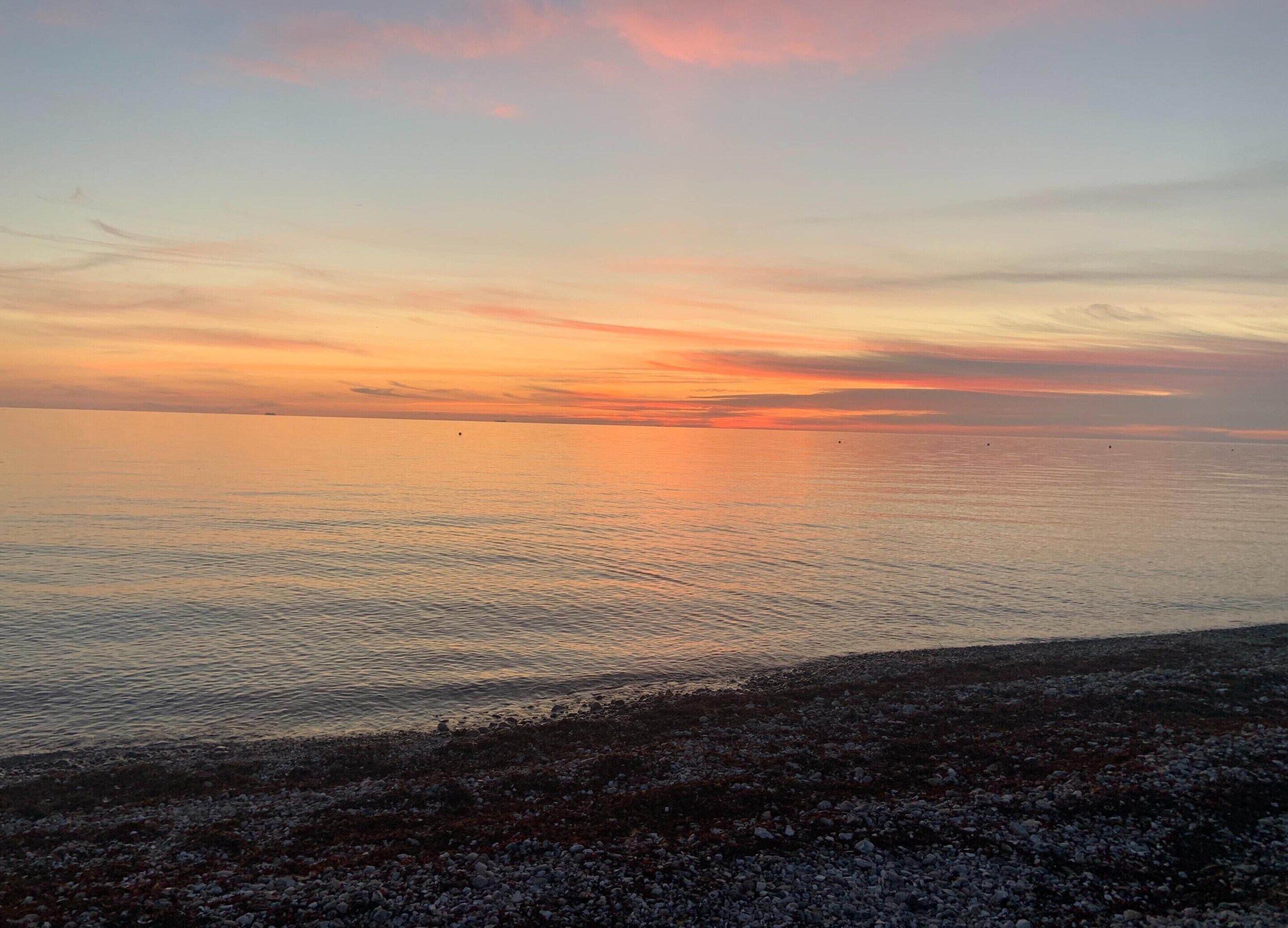 Rantaviiva, jossa on kivikkoista maastoa ja tyyni vesi, johon auringonlasku heijastuu.