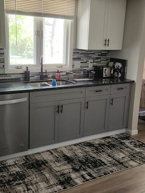 Une cuisine avec des meubles gris, un plan de travail noir, une fenêtre avec des stores et un tapis à motifs.