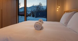 A hotel room with a bed, two rolled towels, a window with a view of snow-covered mountains, and a wooden headboard. - Shion (Kutchan)