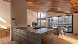 A modern kitchen with a central island, wooden ceiling, and large windows offering a view of trees. - Shion (Kutchan)