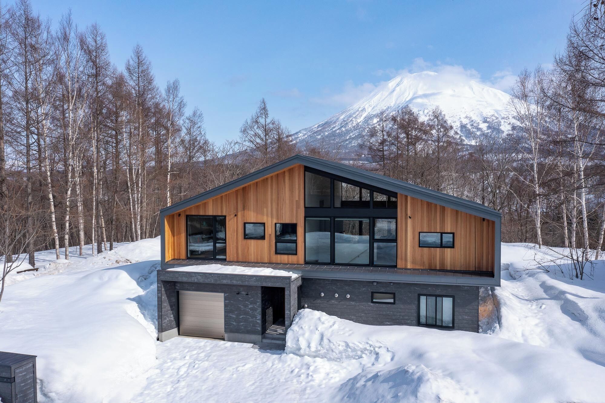 A modern two-story house with large windows, a garage, and a snowy landscape with a mountain in the background.