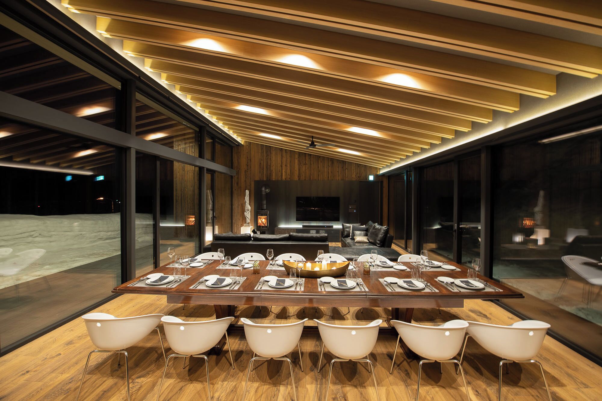 A modern dining area with a long table set for a meal, surrounded by glass walls and a wooden ceiling.