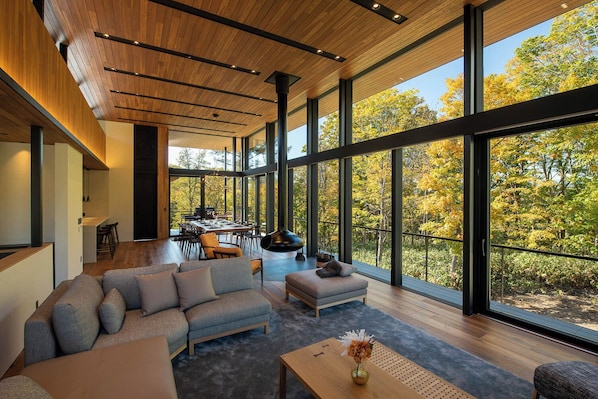 Ein modernes Interieur mit einem großzügigen Essbereich, einer Couch und Blick auf Bäume durch große Fenster.
