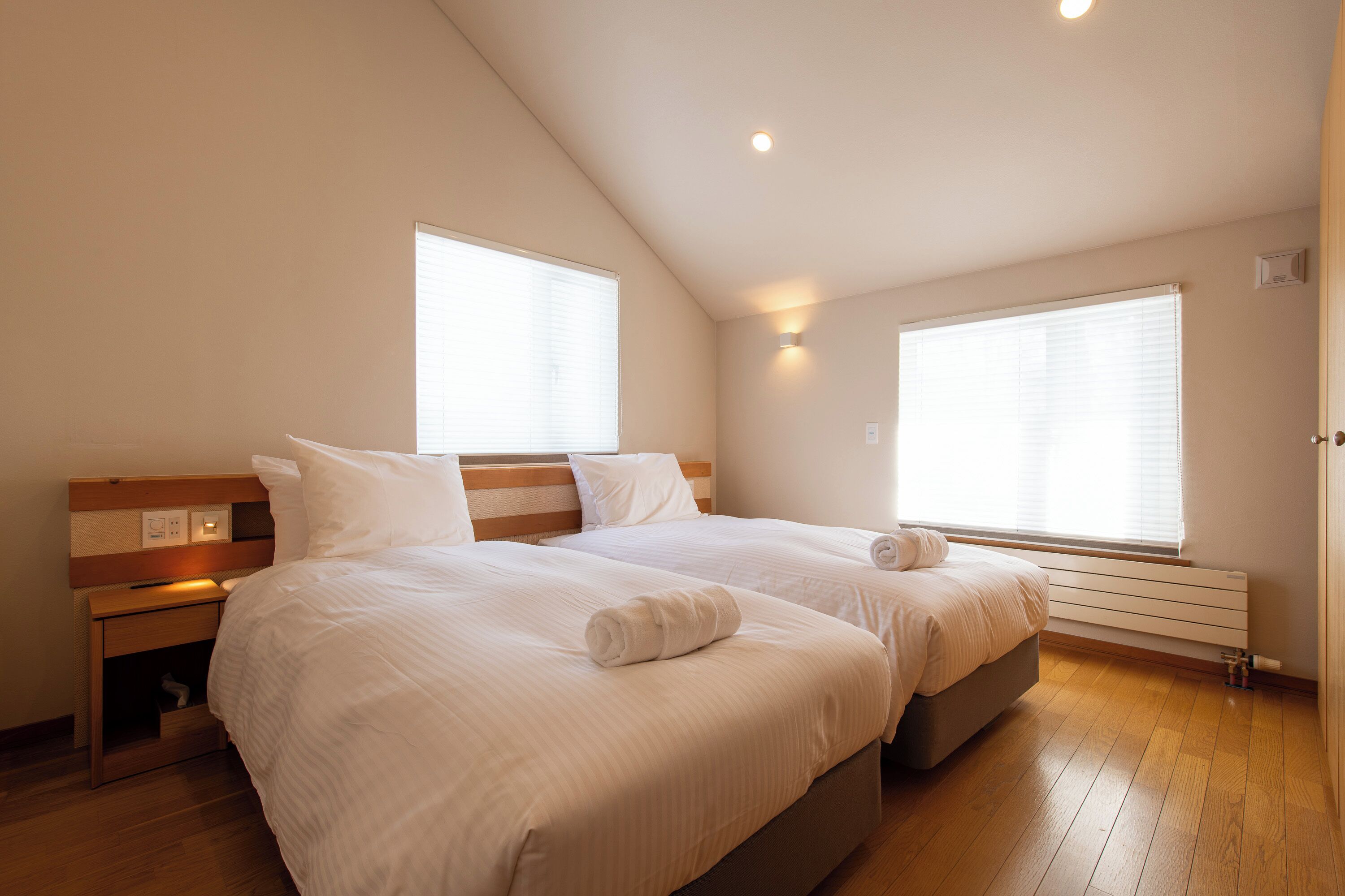 A bedroom with two beds, a nightstand, a wooden floor, and a window with blinds.