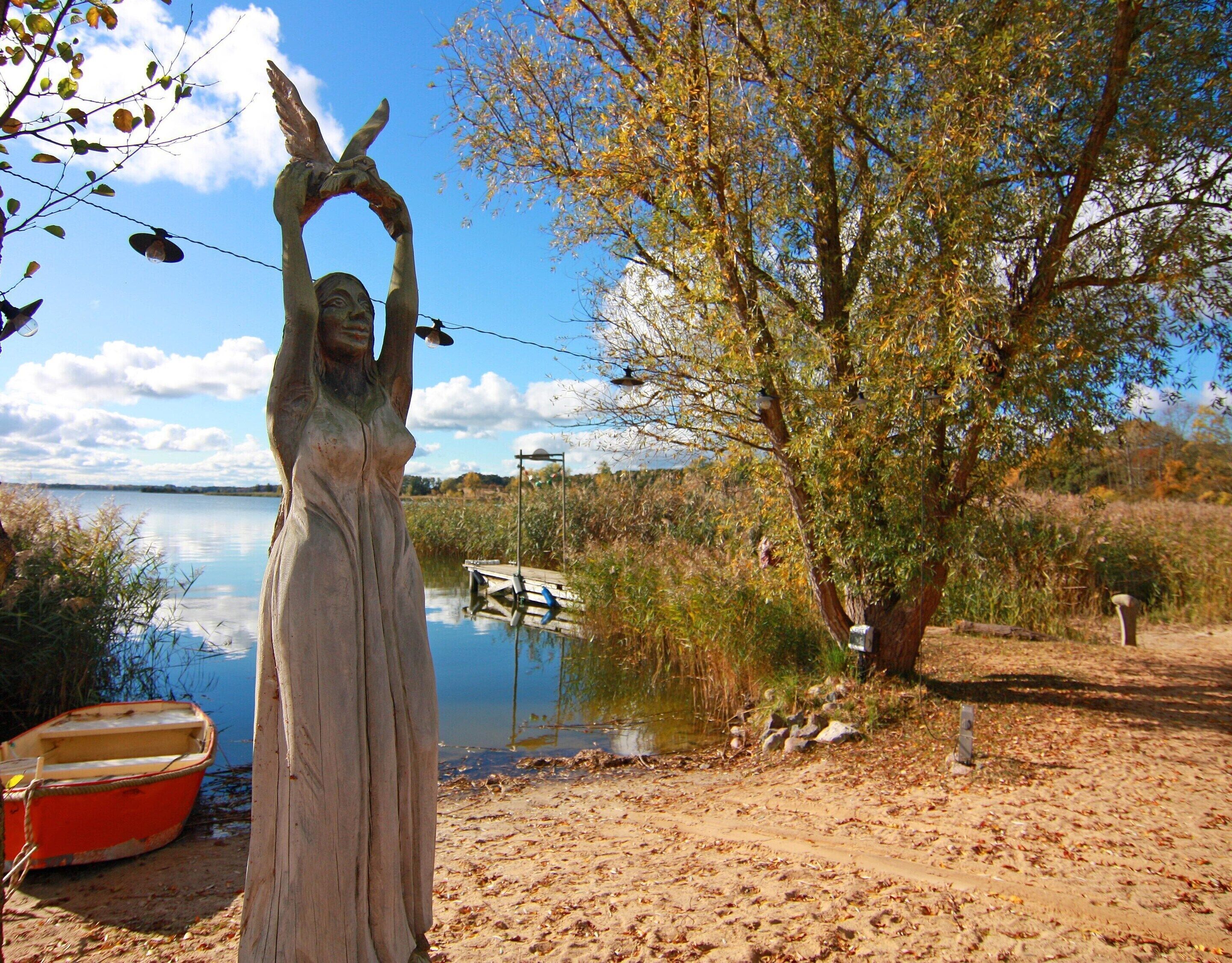 A statue of a woman with raised arms, a boat, and a tree by a body of water.